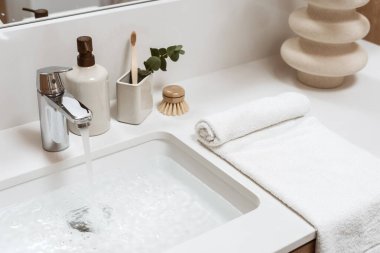 Beyaz lavabo dolu su, metal musluk, şeker kutusu, diş fırçası, bambu fırçası, rulo havlu ve beyaz bir mobilyada seramik vazo.