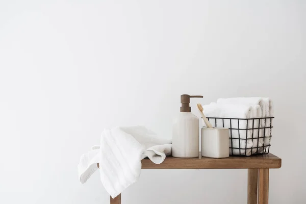 Concept of beauty products for everyday use and skin care routine: wooden bench with folded towels in a metal basket, soap dispenser bottle and a cup with a toothbrush