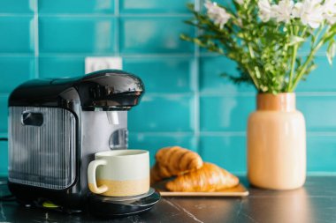 Fincanlı kahve makinesine yakın çekim, ahşap tabakta taze kruvasanlar ve mermer tezgahta çiçekli seramik vazo ve duvarda samimi iç döşeme ve mavi fayanslar var.