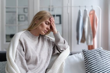 üzgün bir kadın oturma odasında kanepede oturuyor, örgü örüyor, alnına dokunuyor ve migren, zayıflık veya depresyon geçiriyor, hastalık konsepti