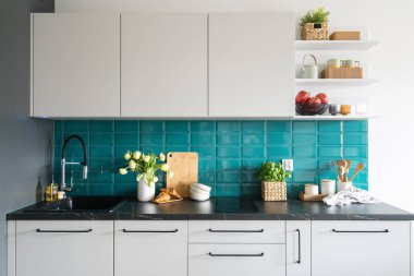 İç dekorasyonlu ev mutfağı, beyaz dolap, çeşmeli lavabo, çiçekli vazo sepette fesleğen, ahşap tabakta taze kruvasan ve duvarda mavi fayans.