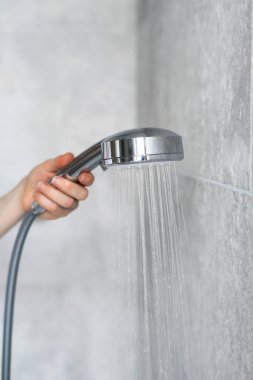 Kadının modern bir duş başlığını temiz banyoda gri fayanslı arka planda tutarken dikey görüntüsü. Su akıyor ve su hortumundan fışkırıyor.
