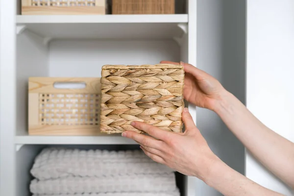 cropped shot of female hands hold wicker storage box and put in on shelf in wardrobe with clothes and clean towels, organized space concept, order inside closet