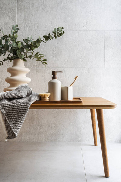 Vertical shot of wooden table with a tray, dispenser bottle, bamboo brush for dry skincare massage, toothbrush in a ceramic cup, towel and vase with a green eucalyptus on tilled white background