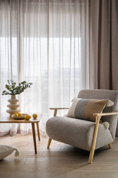 Plants in vase and plate with yellow citrus lemons on wooden table close to comfy soft chair with decorative cushion. Grey armchair perfect addition to light and airy living room in urban apartment