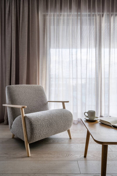 Light living space in modern apartment. Sophistication soft grey armchair against windows with linen curtains. Contemporary furniture and decor in comfortable room. Open book or diary on wooden table