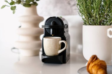 Taze demlenmiş latte, yeni siyah kahve makinesinden evdeki mutfakta seramik fincana dökülüyor. Taze Fransız kruvasanı yakınlarında yeşil bitkileri olan çömlekler beyaz tezgahın üzerinde.
