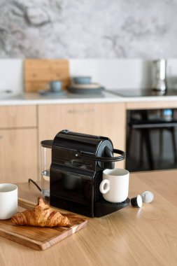 Mutfaktaki ahşap masada beyaz seramik bardak ve espresso makinesinin dikey görüntüsü. Fırınlı kahvaltı tepsisi. Kahvenin yanındaki tabakta taze kruvasan var. İçi bulanık.