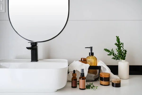 moisturizer cream in glass jar, dispenser bottle, clean towel and vase with plant branch near washbowl on wall background in modern apartment, advertising concept of natural organic cosmetics