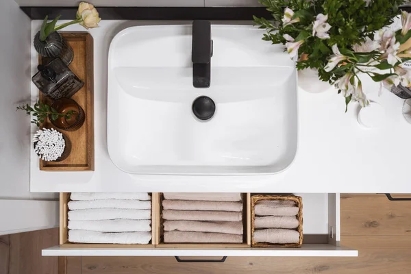 top view on ceramic wash basin with black mixer, flowers in vase, dispenser bottle and glass with ears sticks on wooden tray and open drawer with clean towels in domestic washroom