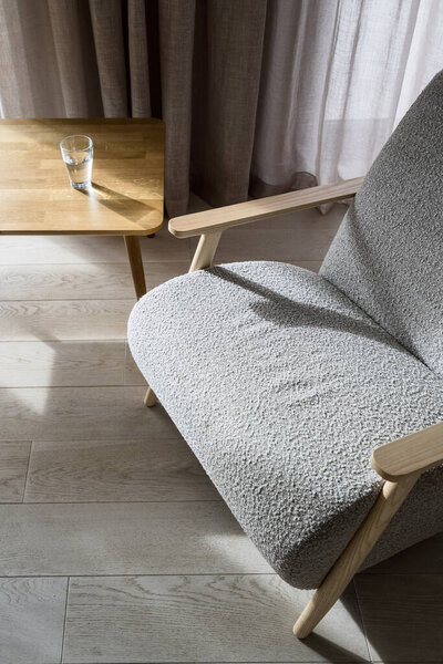 High angle view on empty and stylish armchair with grey upholstery standing near wooden table with glass of drinking water on top. Sophisticated modern apartment. Comfortable furniture in room