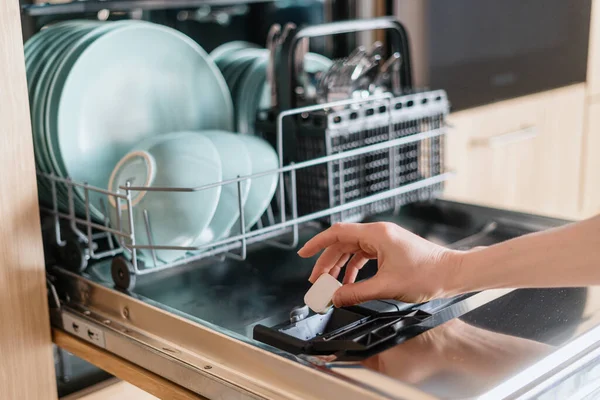 stock image Close up view of female hand inserting dishwasher tablet into open built-in dishwasher machine with utensils inside in modern home kitchen. Household, housekeeping, domestic concept