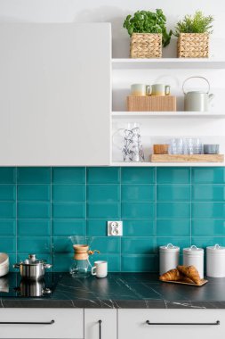 Mutfakta şık bir iç mekan, indüksiyon cam seramik hob, kapaklı metal kavanozlar ve mermer tezgahta ahşap kruvasanlar ve çaydanlık, raflarda fincanlar ve bitkiler.