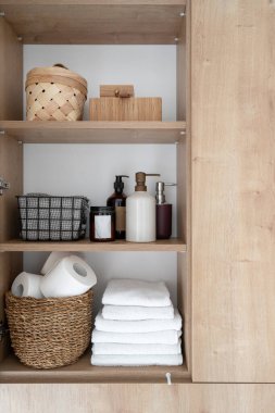 front view on open door of wooden cabinet, wicker basket with toilet paper rolls, neatly folded fresh towels, hygiene products and dispenser bottles on shelves clipart