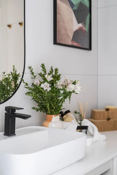 porcelain washbowl with black metal mixer, mirror on tiled wall, vase with bloom flowers and cosmetics products in basket in domestic washroom with elegant interior