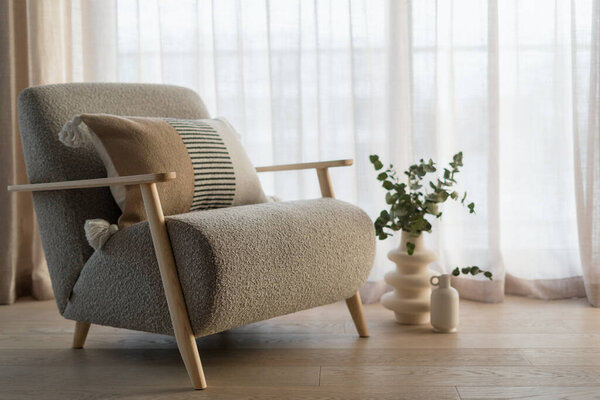 Ergonomic grey armchair and modern vase with eucalyptus plants in stylish and cozy apartment living room. Modern furniture and home decor create a calm and relaxing space