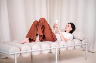 Genç bir kadın elinde akıllı telefonuyla rahat bir şiltenin üzerinde yatıyor. Kadın gündüz yatağında dinleniyor, cep telefonuyla mesaj atıyor. Yumuşak kanepede sohbet için internet uygulamalı cep telefonu kullanan bir kız.