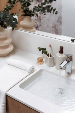 Lavabo dolu suyun üzerine krom musluk, kişisel aksesuarlara yakın. Sabun makinesi şişesi, fincanda ahşap diş fırçası bambu masaj fırçası, yeşil okaliptüslü havlu ve vazo.