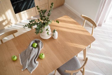 Taze elmaların üst görüntüsü, bulaşık havlusu ve okaliptüs dallı seramik vazo modern mobilyaları ve iç dekorasyonu olan şık mutfağın ahşap masa üstü.