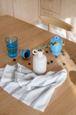 Pamuk bulaşık havlusu, okaliptüs dallı seramik vazo, ahşap masa üstünde bir bardak su ve taze yaban mersini, yüksek açılı çekim.