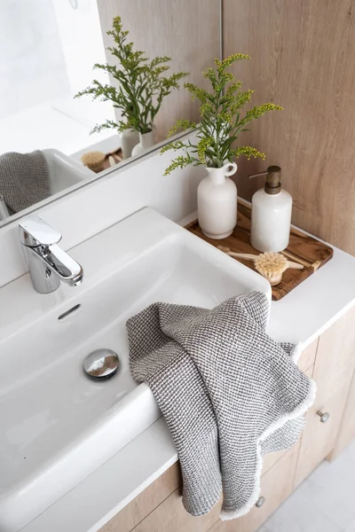 stock image towel on porcelain washbasin, chrome water tap, massage brush, vase with plant and dispenser bottle with soap on wooden tray on countertop in domestic washroom, high angle shot