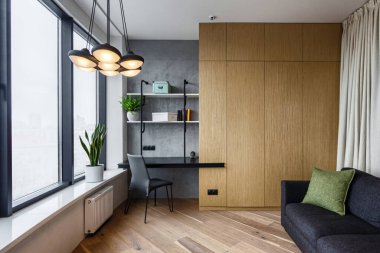Ahşap gardırobu ve gri rahat koltuğu olan bir oda. Ofis masası ve pencerenin yanındaki sandalye. Modern iç mekan konsepti şık bir dairede