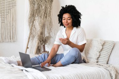 Gülümseyen Afrikalı Amerikalı serbest çalışan kadın sosyal medyada sohbet etmek için akıllı telefon kullanıyor ve evde yatakta otururken laptopta bilgi tarıyor. uzak çalışma konsepti
