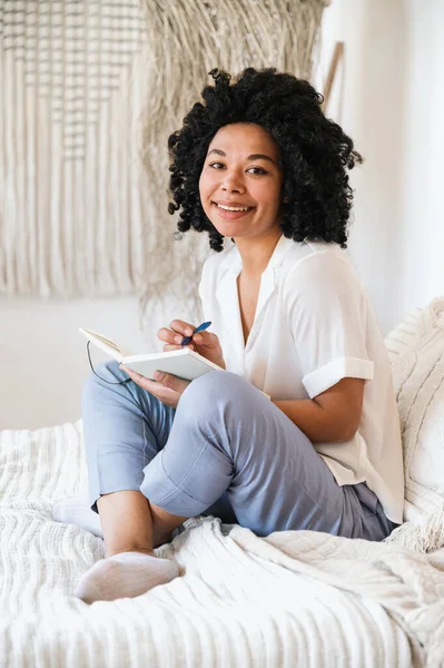 stock image happy afro american woman freelancer taking notes in notebook planner while sitting in bed at home. smiling woman write inspiration ideas in journal diary
