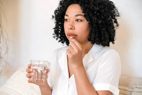 stock image african american woman hold glas of water and taking medication from head ache or stomach pain. young female suffering from depression and has sedative pill. disease treatment concept