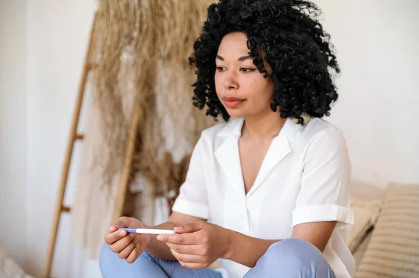 sad disappointed afro american woman hold pregnancy test while sitting on couch alone at home. young female feeling troubled by result, has health problems or infertility