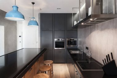 Siyah tezgahlı mutfak adası, bar taburesi, modern dolap, indüksiyon sobası, elektrikli fırın. Stüdyoda musluk ve mavi tavan lambalarıyla lavabo.