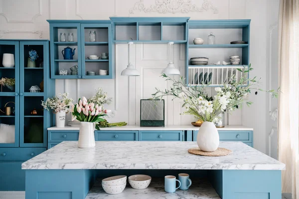 Marble countertop with vases and flowers in provence style apartment. Kitchen island and dining table with tableware. Blue furniture and white walls in classic interior design room.