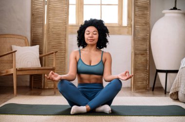 Spor giyim bölümündeki rahat Afro-Amerikan kadın gözlerini kapattı, yere oturdu ve yogada lotus asana pratiği yaptı. Padmasana pozu. Evde meditasyon yapıyordum. Ruh sağlığı hizmetleri kavramları