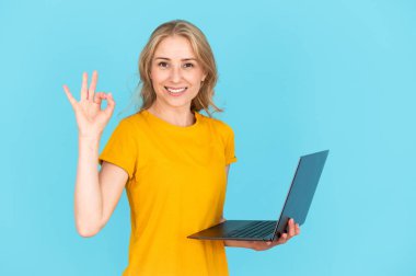 Modern dizüstü bilgisayarı tutan neşeli bir kadının portresi, ok işaretini gösteriyor, mavi fotokopi arkaplanda tek başına duruyor. Kadın gülümsemesi, iyi kalitede internet bağlantısı, web sağlayıcısı