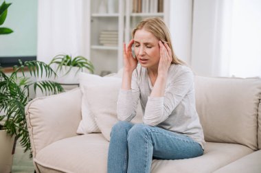 Bitkin düşmüş genç bir kadın şakaklara dokunuyor, bej koltukta tek başına oturuyor, şiddetli baş ağrısı ya da migrenden muzdarip. Sağlık sorunları, kötü refah