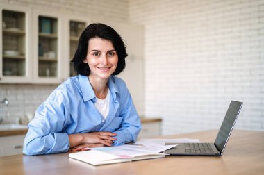 Masada oturan gülümseyen genç bir kadın, kameraya bakıyor ve internetten ders çalışıyor. Modern dizüstü bilgisayar, defter ve belgeler yemek odasındaki ahşap masada. Uzaktan öğrenme, ev ofisinde çalışma