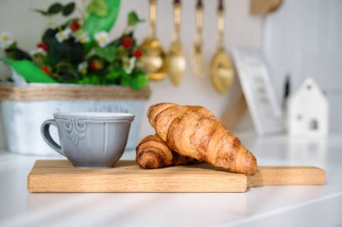 Fransız kruvasan ve fincana yakın çekim. Mutfak arka planında ahşap kesme tahtasının üzerinde kahve var. Evdeki masada taze pasta ve çay var. Nefis kahvaltı konsepti