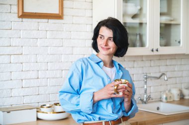 Gülümseyen bir kadının portresi. Mutfakta duruyor, başka tarafa bakıyor, elinde seramik bardakla kahve içiyor. Rahat bir evde mükemmel bir sabah. Bir kadın yeni hafta sonu hakkında düşünüyor ve hayal kuruyor..