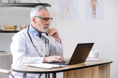 Gözlüklü orta yaşlı bir doktor. Klinik dolabında dizüstü bilgisayarla oturmuş hasta kayıtlarını araştırıyor. Randevu için hazırlanıyor. sağlık hizmeti kavramı