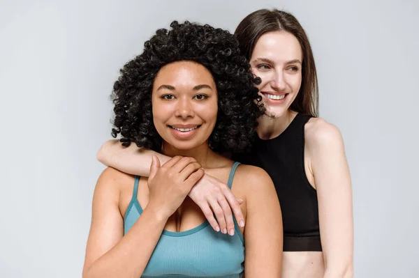 Diversity and different ethnicity portrait concepts. Fitness girls in sportswear hug, looking at camera and smiling together isolated on gray background. Leisure and physical activity