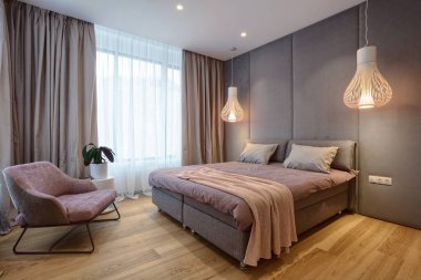 Çift kişilik yatağı ve gri ve yumuşak duvarlı rahat koltuğu olan zarif bir yatak odası. Modern iç mekanda şık detaylar kavramı. Tekstil, yastık, battaniye ve perdeli ev dekoru.