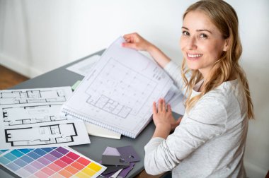 High angle view on young smiling female architect working with plans, documents, technical drawings, blueprint drafts and color palettes at workplace. Good ideas for arrangement new house clipart