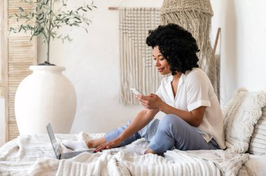 Neşeli Afro-Amerikan kadın defter üzerinde çalışıyor ve modern akıllı telefonundaki internet uygulamalarını kontrol ediyor. Yatakta rahat bir şekilde oturan serbest bir kadın blog için yazı yazıyor. Ev kavramlarından uzak çalışma