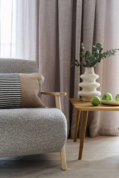 Soft grey couch or comfortable armchair standing near wooden table in light modern apartment. Design furniture and home decor in stylish and contemporary living room interior