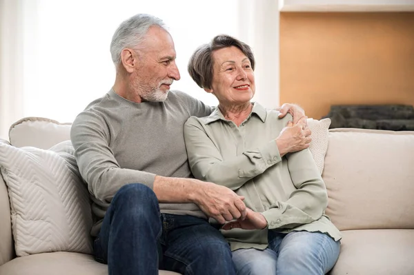 elderly couple sitting on couch in living room watching TV hugging and spending time together. senior woman and man resting on sofa in living room. happy retirement couple. weekend at home concept