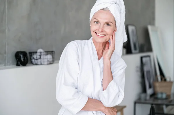 Smiling mature woman in robe and towel on head after taking shower in bathroom at home. Washing hair with shampoo without parabens and using repair mask.