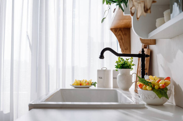side view on kitchen sink and black faucet in vintage style. bowl with ripe grape, green fresh parsley in jug and metal food containers on white countertop in bright room