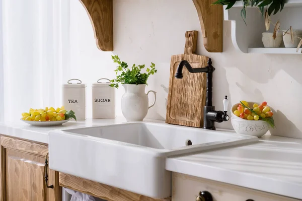 decor at home kitchen, fresh parsley in jug, grape on plate, tea and sugar in metal food canisters and cutting board on white countertop near ceramic sink and water tap in retro style