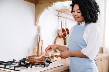 Gülümseyen Afro-Amerikan temizlikçi mavi önlüklü aile yemeği pişiriyor. Yemeği paslanmaz tavada karıştıran kadın, sabahın köründe rahat bir mutfakta duran kadın.