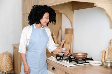 Mavi önlüklü gülümseyen ev hanımı elinde tavayla yemek pişiriyor. İskandinav mutfağındaki ahşap mobilyalar ve aksesuarlar ahşap mobilyalar ve beyaz duvarlar..